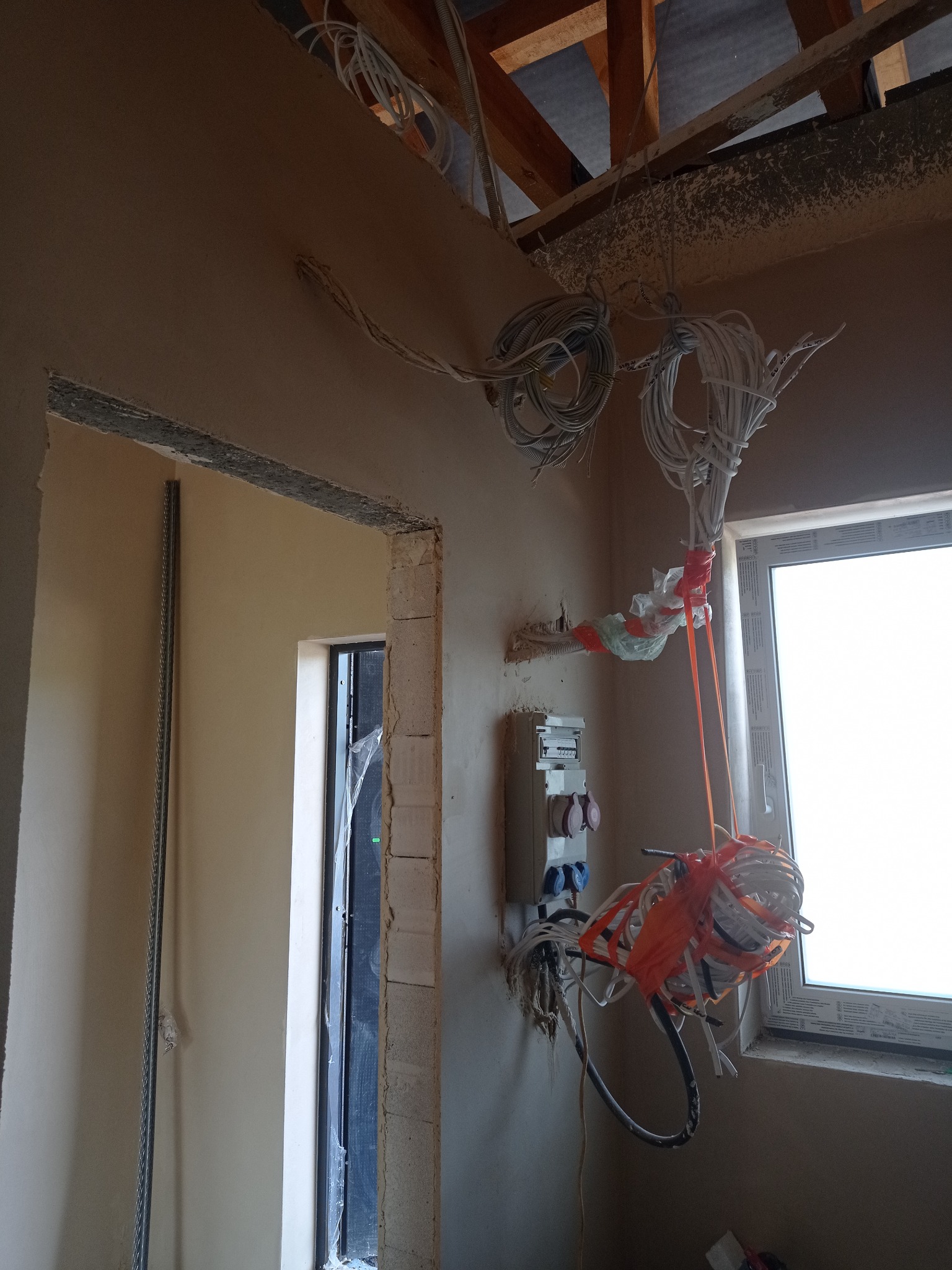 Świeżo otynkowane ściany w surowym stanie z widoczną instalacją elektryczną, kable zwisają z sufitu. W tle okno i fragment drewnianej konstrukcji dachu.