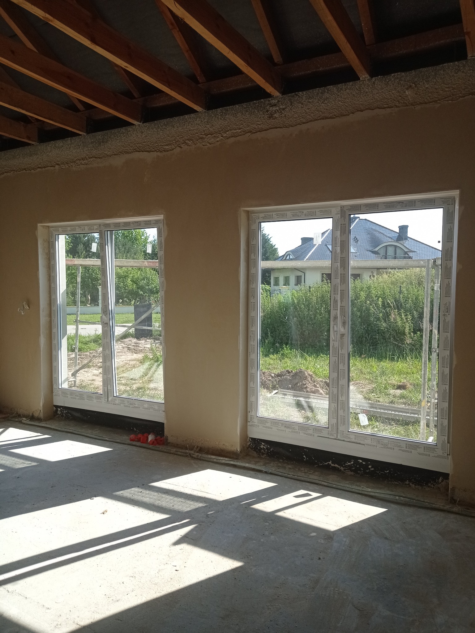 Wnętrze w stanie surowym z dwiema parami białych okien PCV, świeżo otynkowane ściany z widoczną warstwą izolacji pod sufitem i surowym drewnianym stropem, na zewnątrz widok na budynek i zieleń.