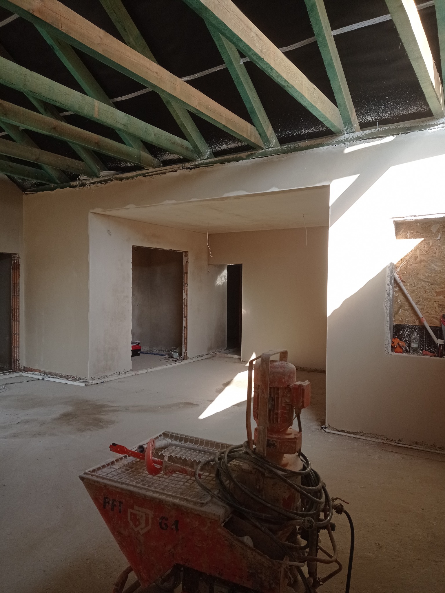 Wnętrze w trakcie prac wykończeniowych: ściany pokryte świeżym tynkiem, widoczna maszyna tynkarska FFT G4, surowe drewniane belki stropowe, okno z prowizorycznym zabezpieczeniem z płyty OSB.