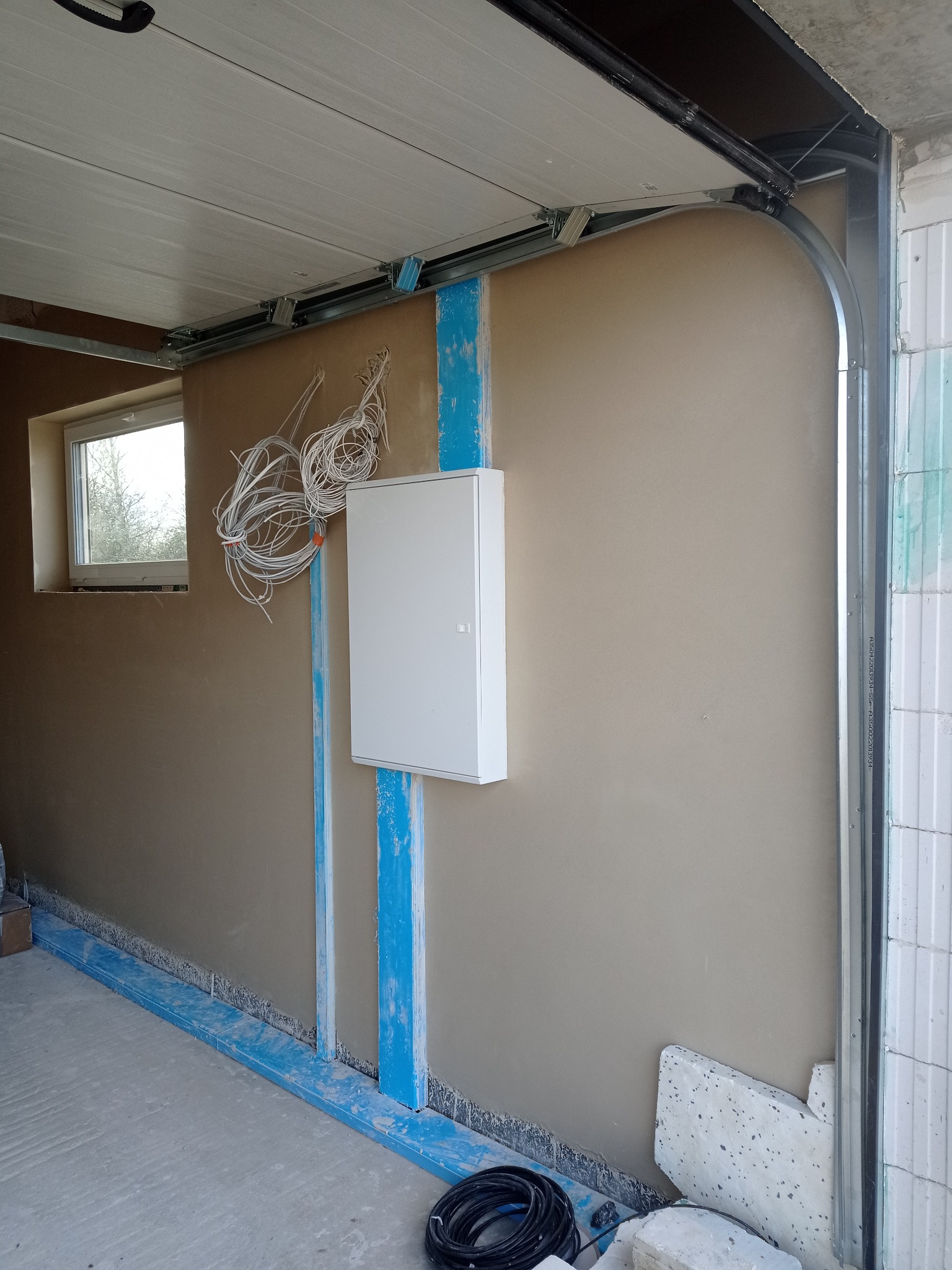 Świeżo położony beżowy tynk na ścianie garażu, widoczna puszka elektryczna z wystającymi przewodami, okno oraz elementy konstrukcyjne bramy garażowej.
