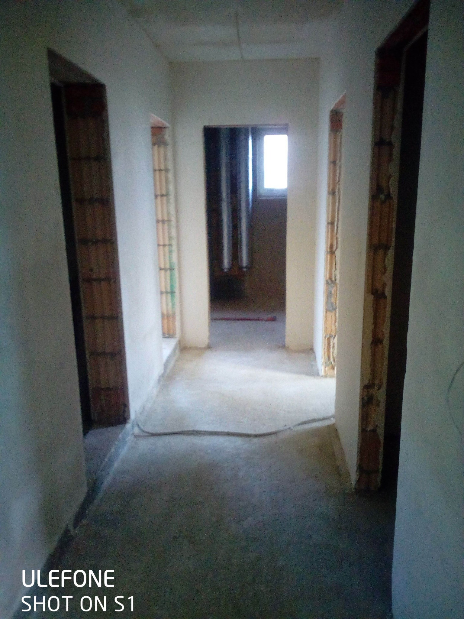 Korytarz w surowym stanie po nałożeniu wstępnej warstwy tynku, widoczne ościeżnice bez drzwi i niedokończone ściany, światło dzienne wpadające przez okno na końcu korytarza.