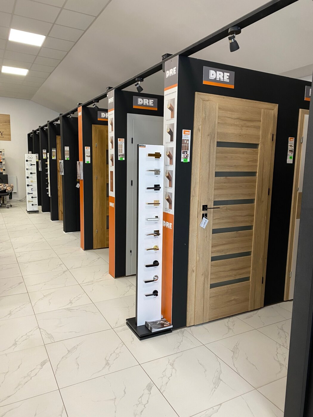 Ekspozycja różnych modeli drzwi wewnętrznych marki DRE w salonie sprzedaży, widoczne próbki klamek i oświetlenie punktowe.