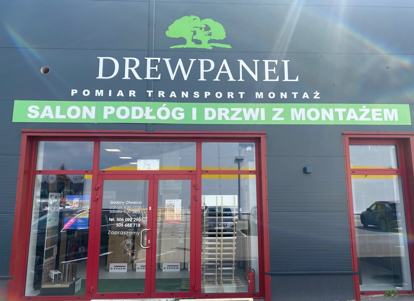 Fasada budynku salonu 'Drewpanel' z logo i napisem 'Salon podłóg i drzwi z montażem' nad czerwonymi witrynami wystawowymi.