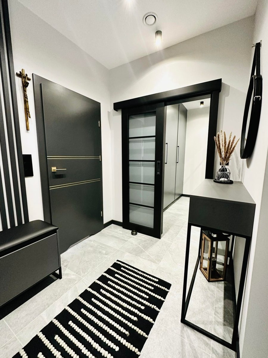 Nowoczesny przedpokój w odcieniach szarości i czerni z przesuwnymi drzwiami szklanymi, dywanikiem w geometryczne wzory i minimalistyczną konsolą z dekoracją.
