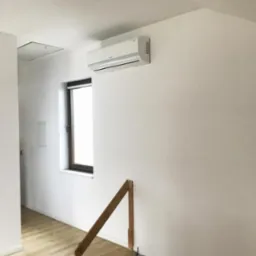 Biały klimatyzator ścienny zamontowany w jasnym pomieszczeniu z drewnianą balustradą schodów i oknem.