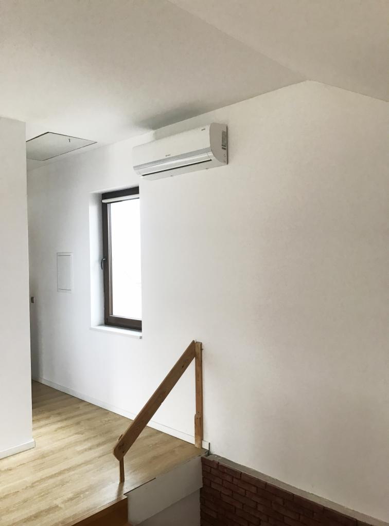 Biały klimatyzator ścienny zamontowany w jasnym pomieszczeniu z drewnianą balustradą schodów i oknem.