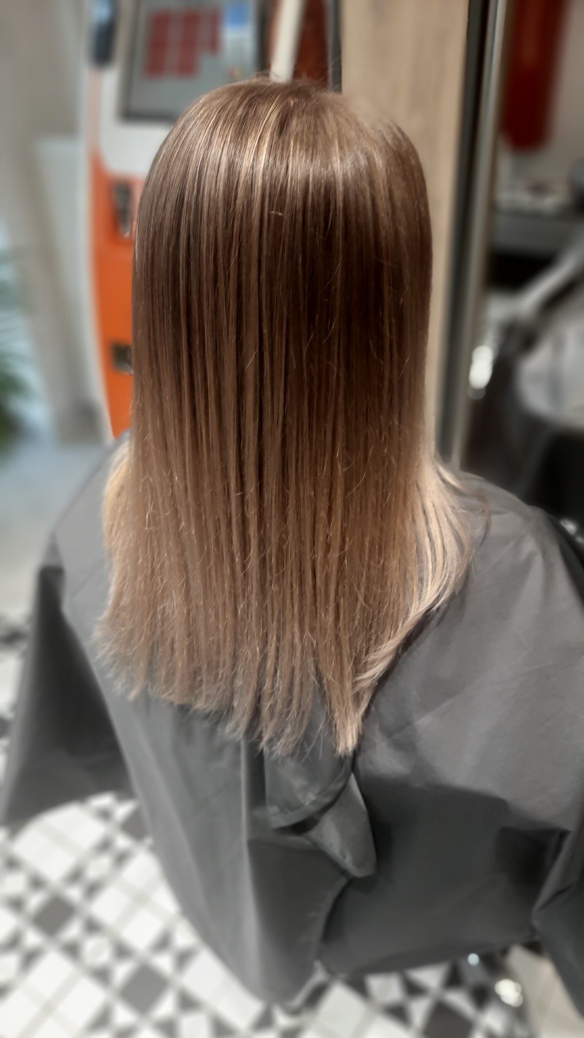 Włosy po koloryzacji sombre, widoczne od tyłu, z jaśniejszymi końcówkami i ciemniejszymi odrostami, założony fartuch ochronny, podłoga w biało-szarą szachownicę.