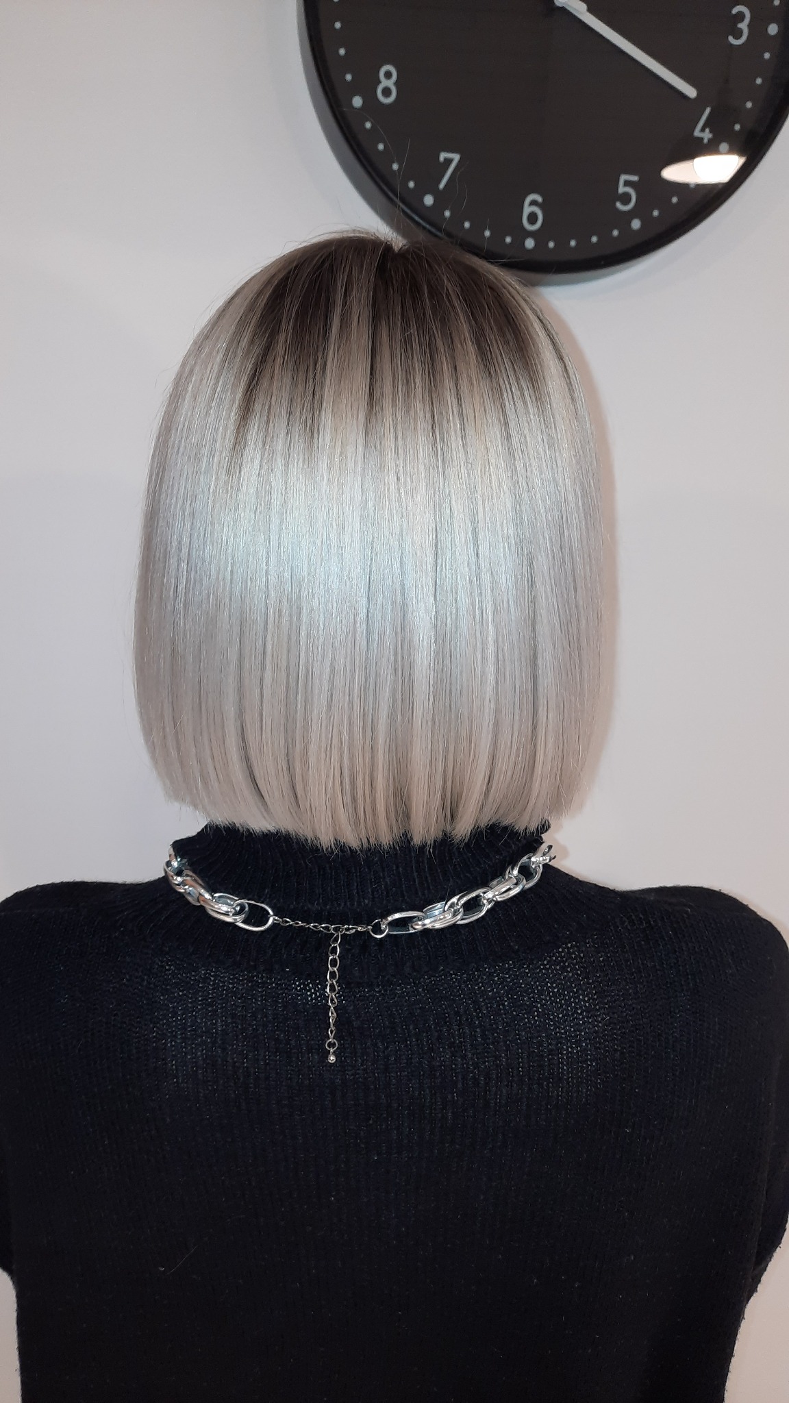 Krótkie, platynowe włosy z ciemniejszym odrostem, widok z tyłu, ozdobny srebrny łańcuch na szyi, czarny sweter, zegar na ścianie w tle.
