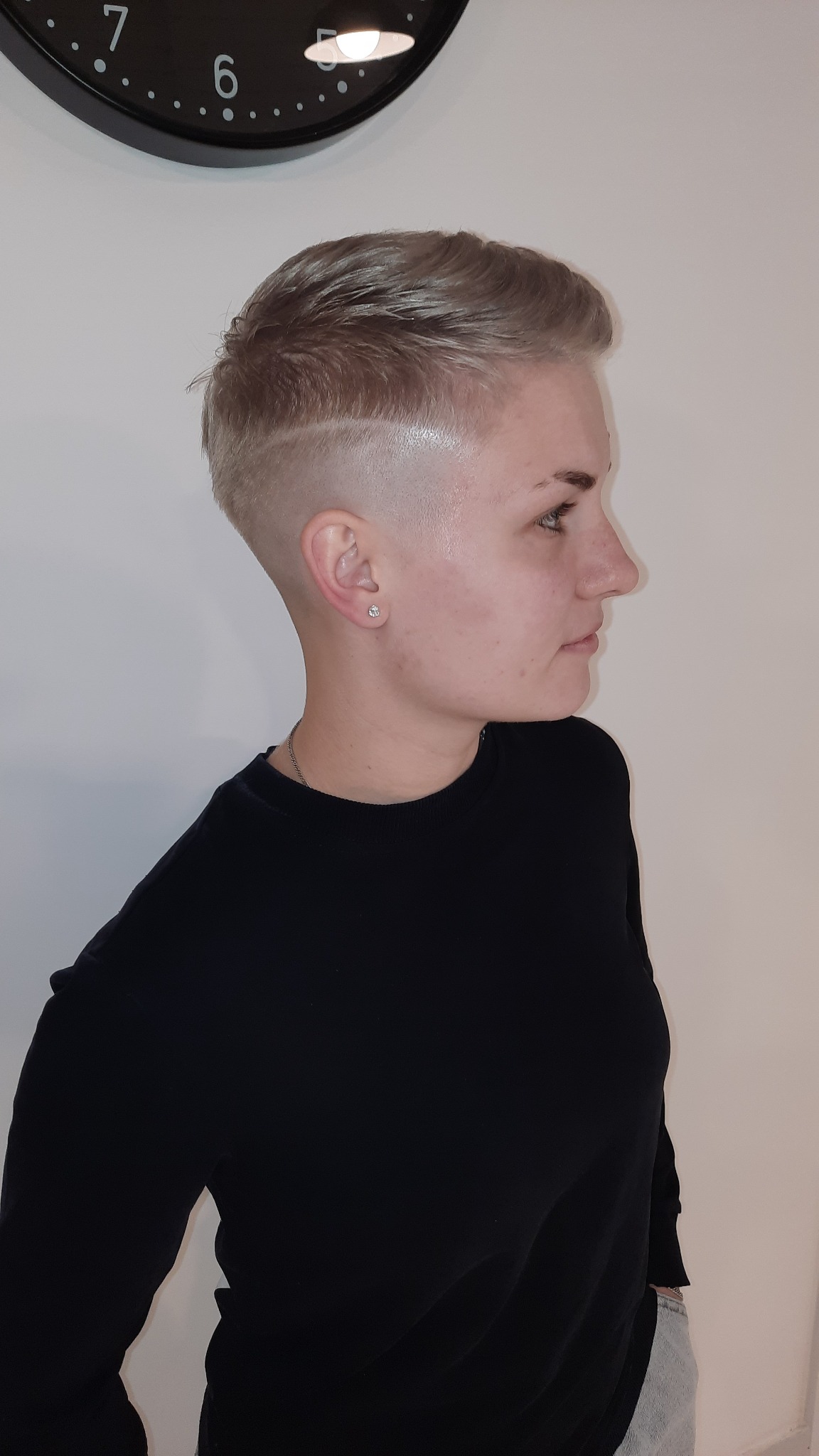 Krótkie, blond włosy z wygolonym bokiem i delikatnym wzorkiem, widok z profilu, subtelny makijaż, czarna bluzka.