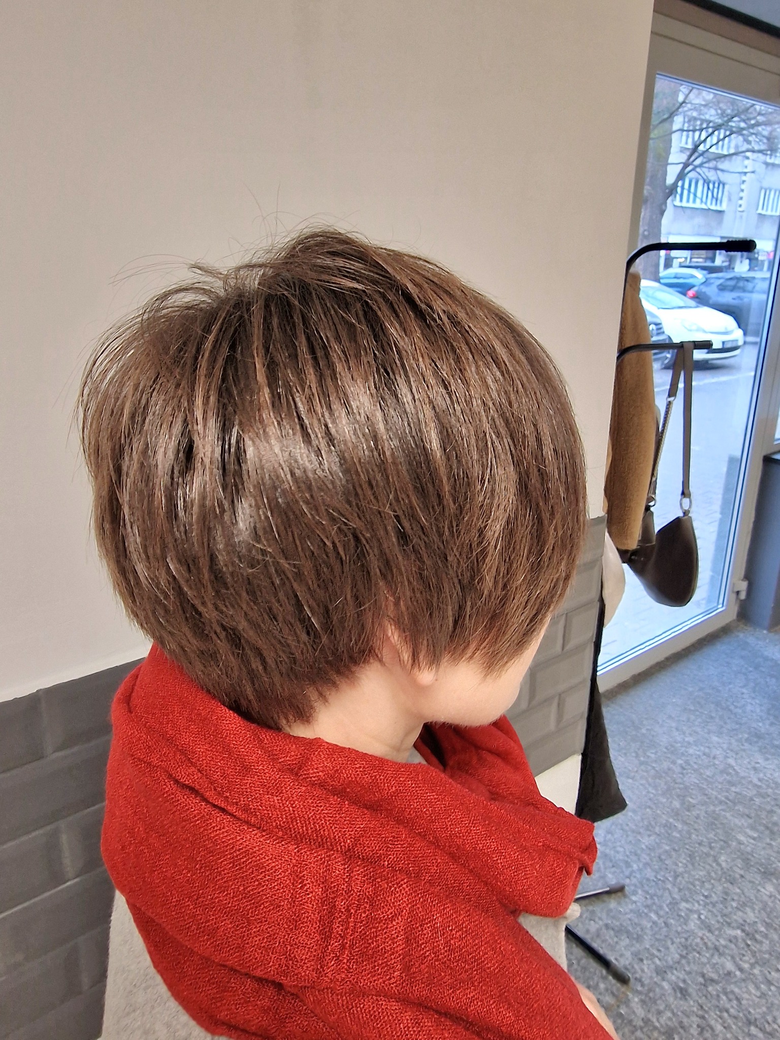 Krótkie, brązowe włosy po strzyżeniu, widok z tyłu, z czerwonym szalem na szyi, na tle okna z widokiem na ulicę.