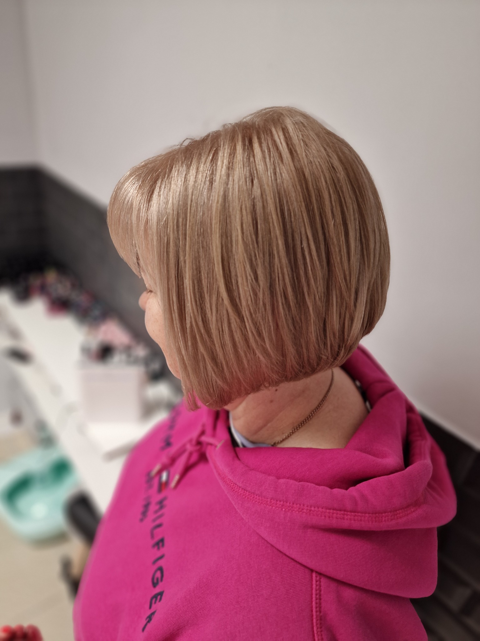 Krótkie, blond włosy z delikatnym cieniowaniem, widoczne z tyłu, na kobiecie w różowej bluzie z kapturem, delikatny makijaż, w tle salon.