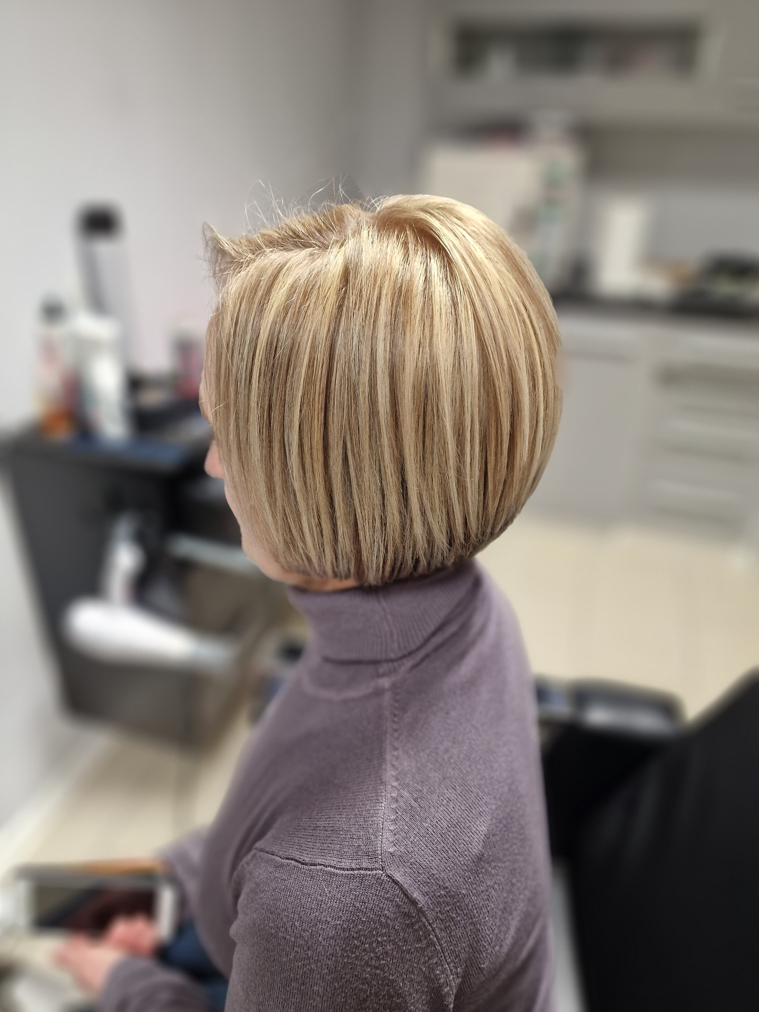 Krótkie, blond włosy z delikatnym cieniowaniem, widoczne z tyłu głowy kobiety w fioletowym golfie, w tle rozmyte wyposażenie salonu.