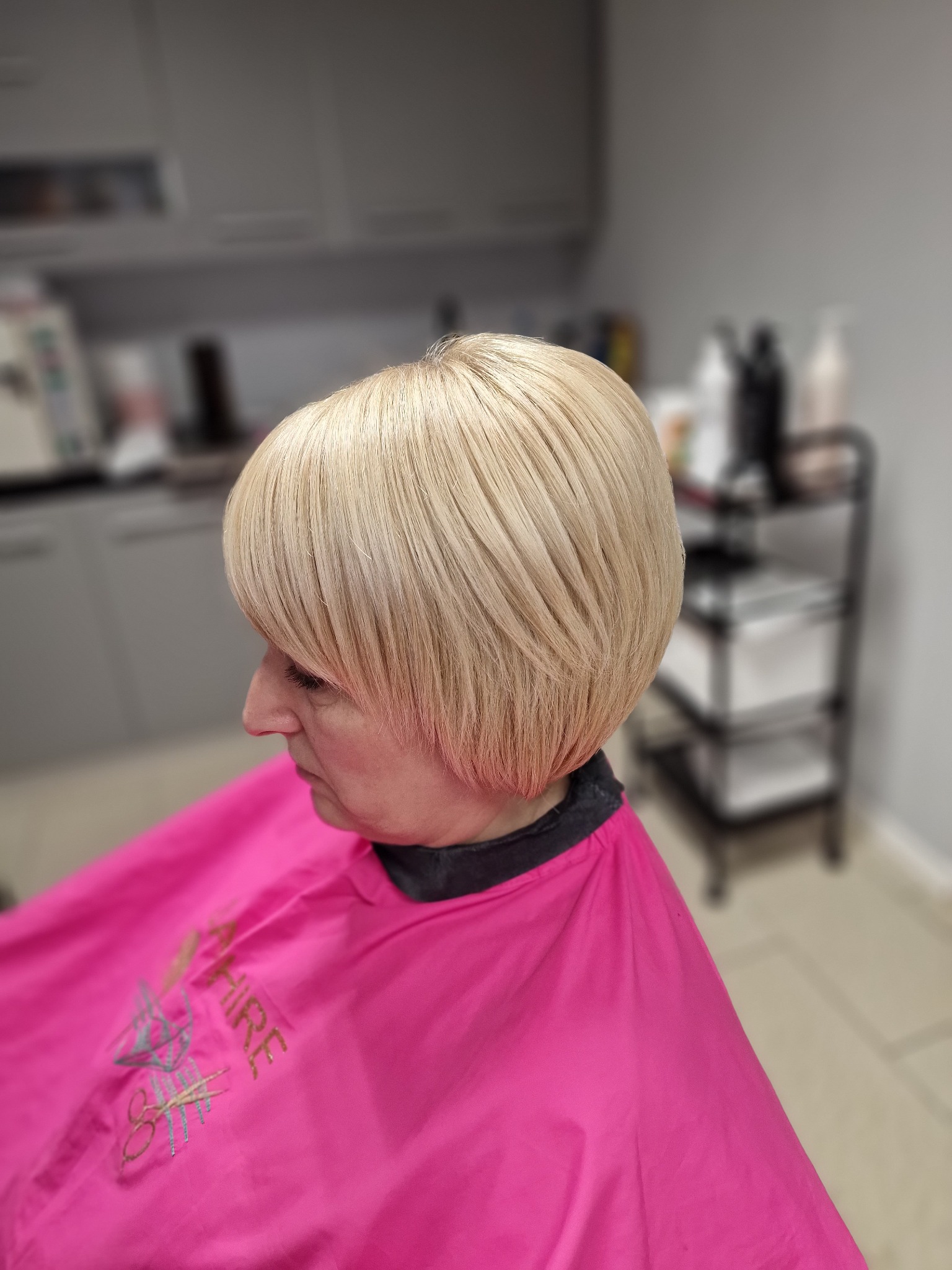 Krótka, blond fryzura z grzywką na kobiecie w różowej pelerynie ochronnej, widok z profilu w salonie.