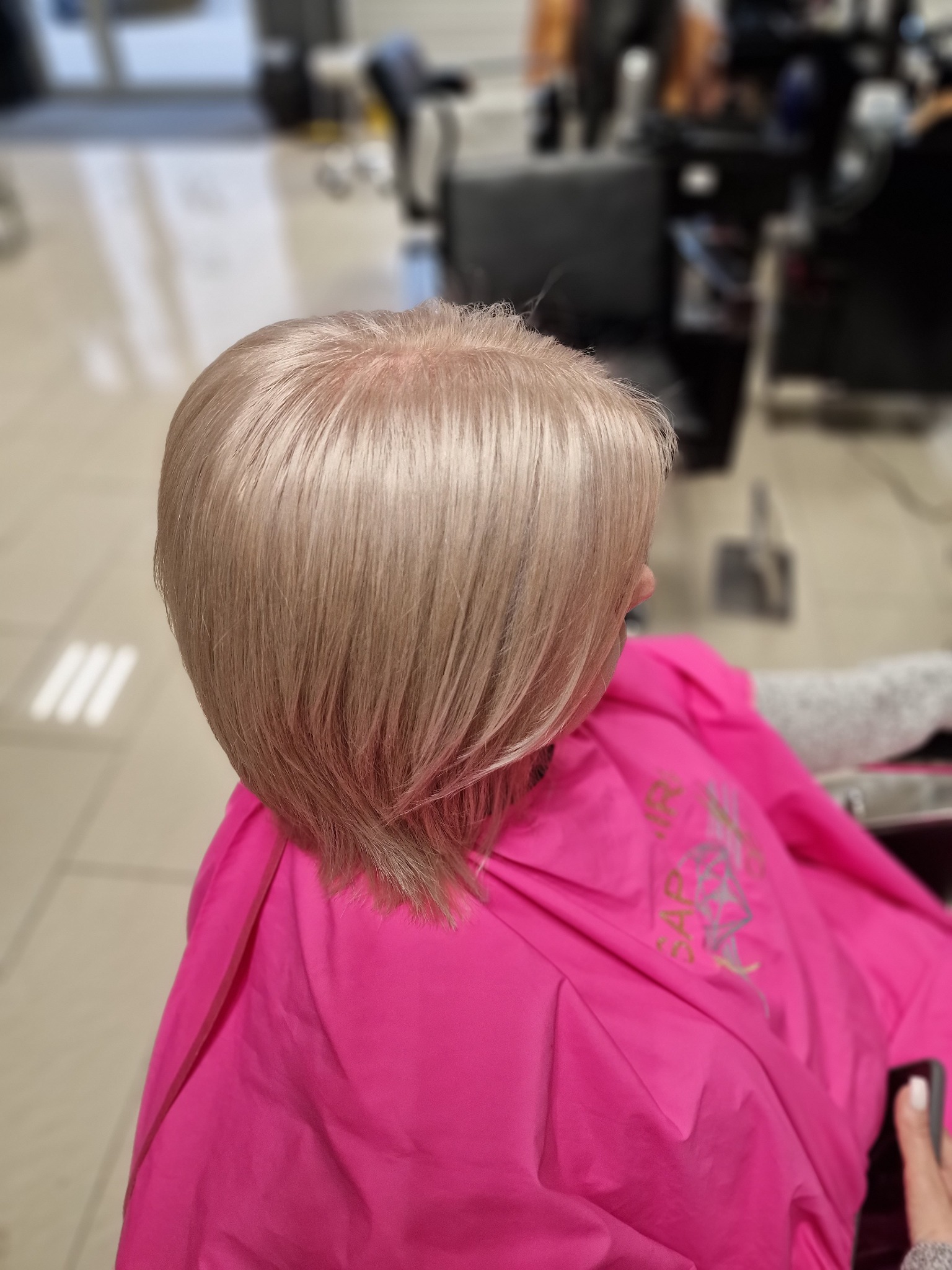 Świeżo ostrzyżone, platynowe blond włosy, widok z tyłu, peleryna ochronna w kolorze fuksji, salon fryzjerski w tle.