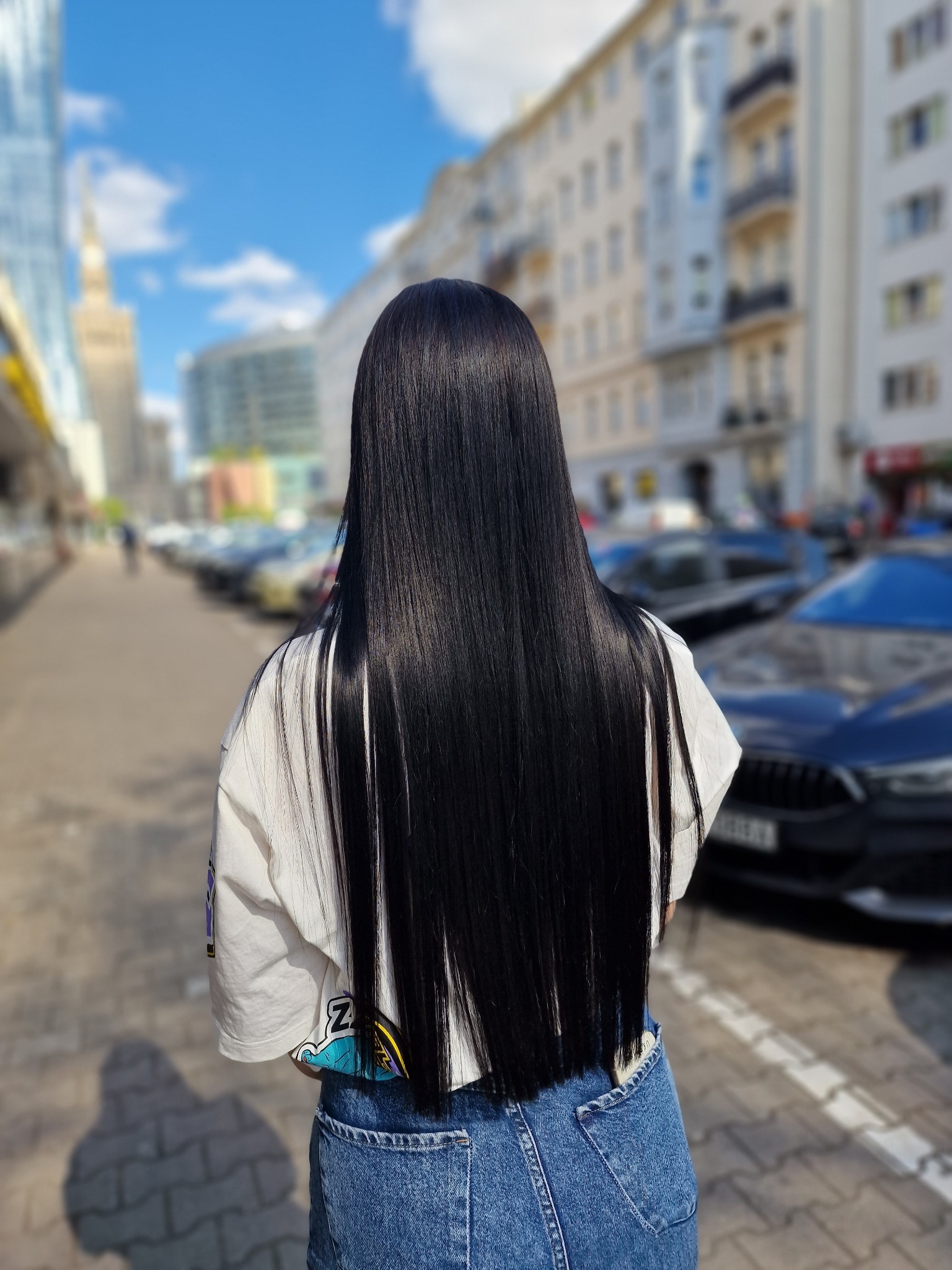 Długie, proste, czarne włosy kobiety, widok z tyłu na tle miejskiej ulicy z budynkami i samochodami w słoneczny dzień.