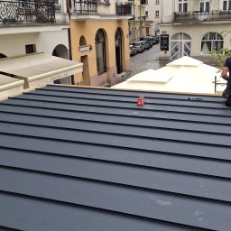 USŁUGI BLACHARSKO-DEKARSKIE PIOTR SZYMCZAK - Nowy, grafitowy dach z blachy na tle miejskiej zabudowy. Widoczne narzędzia i fragment sylwetki dekarza. Dach z wyraźną fakturą, w trakcie montażu.