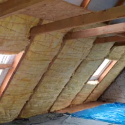 Ocieplenie poddasza wełną mineralną widoczne między krokwiami, zabezpieczone sznurkiem. Fragment okna dachowego i podłogi z folią ochronną.