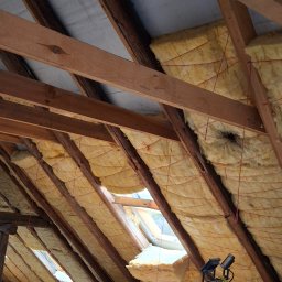 USŁUGI BLACHARSKO-DEKARSKIE PIOTR SZYMCZAK - Poddasze z odsłoniętą konstrukcją drewnianą i izolacją z wełny mineralnej, zabezpieczoną sznurkiem, widoczne okno dachowe i fragment oświetlenia.