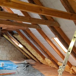 USŁUGI BLACHARSKO-DEKARSKIE PIOTR SZYMCZAK - Poddasze z drewnianą konstrukcją dachu, ocieplone wełną mineralną, zabezpieczone folią. Widoczne okno dachowe i drabina. Kamienna ściana szczytowa w tle.