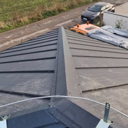 USŁUGI BLACHARSKO-DEKARSKIE PIOTR SZYMCZAK - Szary dach z blachy na budynku jednorodzinnym, widok z góry, z elementami montażowymi i zabezpieczeniami. W tle pole i zabudowania.