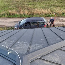 USŁUGI BLACHARSKO-DEKARSKIE PIOTR SZYMCZAK - Szary dach z blachy na rąbek stojący, zabezpieczony systemem odśnieżania. W tle auto i dekarz z narzędziem na tle pola i drogi.