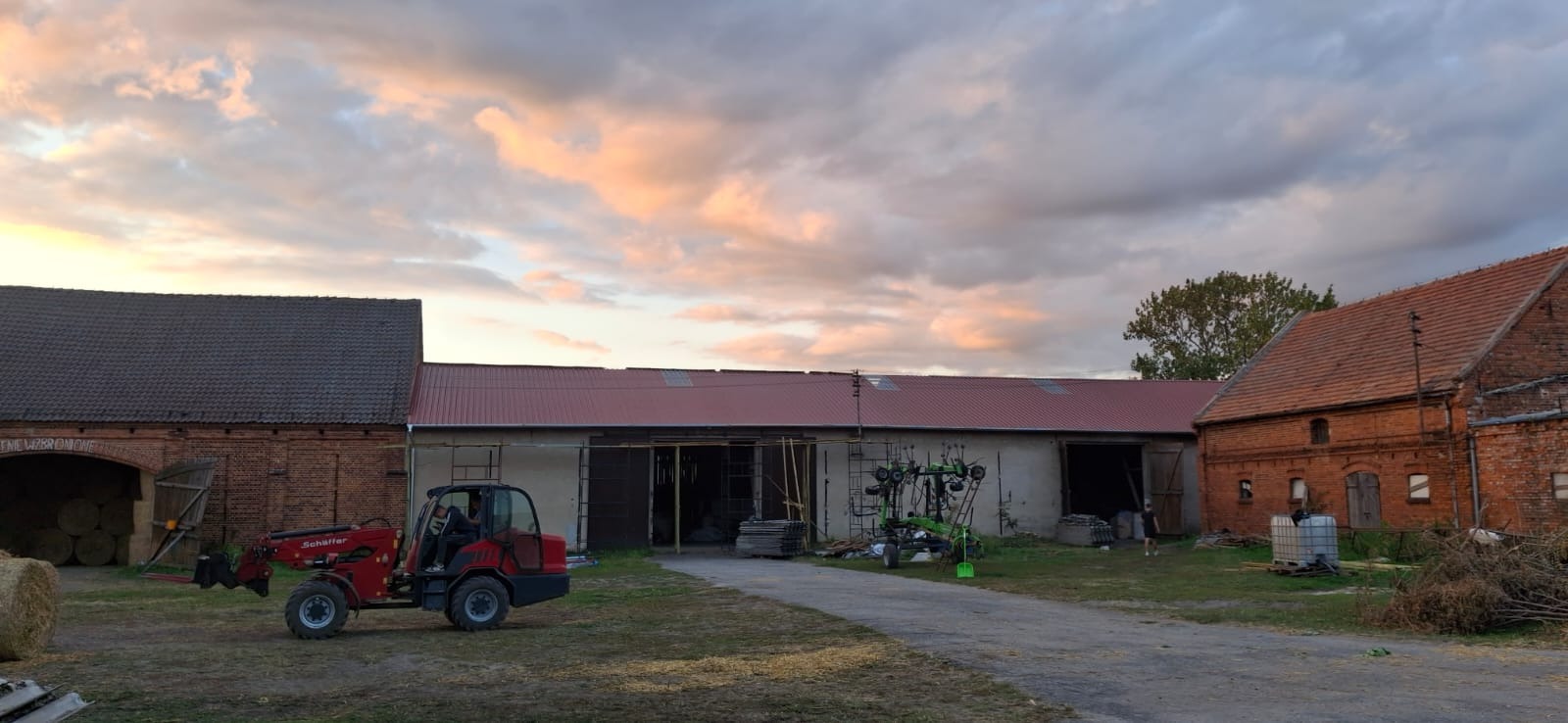 Czerwony dach z blachy trapezowej na budynku gospodarczym w wiejskim krajobrazie o zmierzchu, z widocznym traktorem i stodołą w tle.