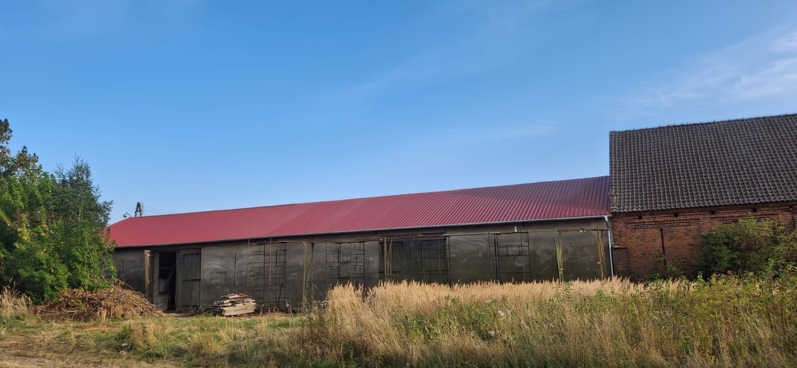 Budynek gospodarczy z nowym, czerwonym dachem z blachy trapezowej. Widoczne metalowe konstrukcje i trawa na pierwszym planie. Drugi budynek z cegły z ciemnym dachem w tle.