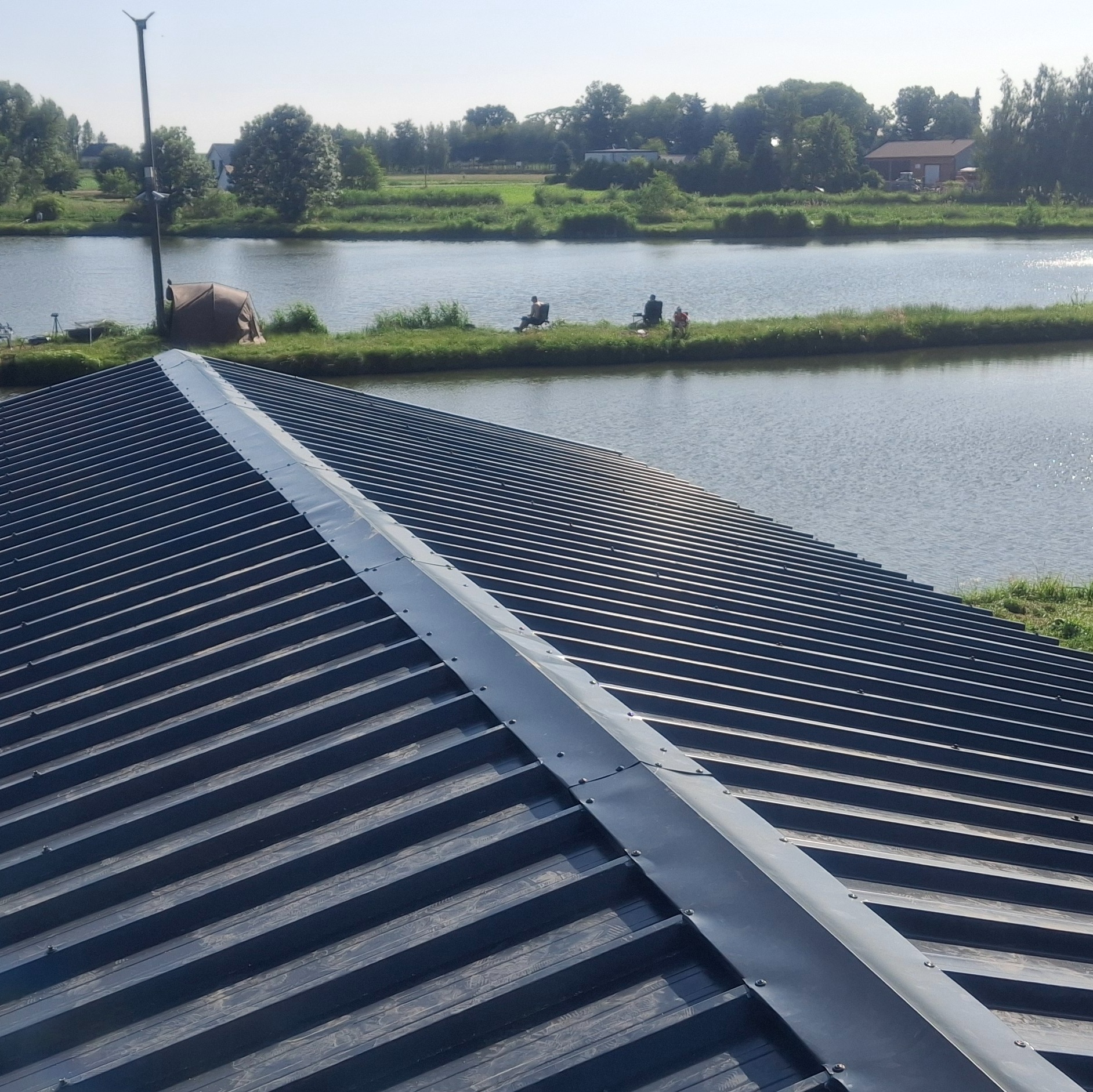 Nowe, grafitowe krycie dachu z blachy trapezowej, widok z góry. Na drugim planie malowniczy krajobraz z jeziorem i wędkarzami. Elementy obróbki blacharskiej.