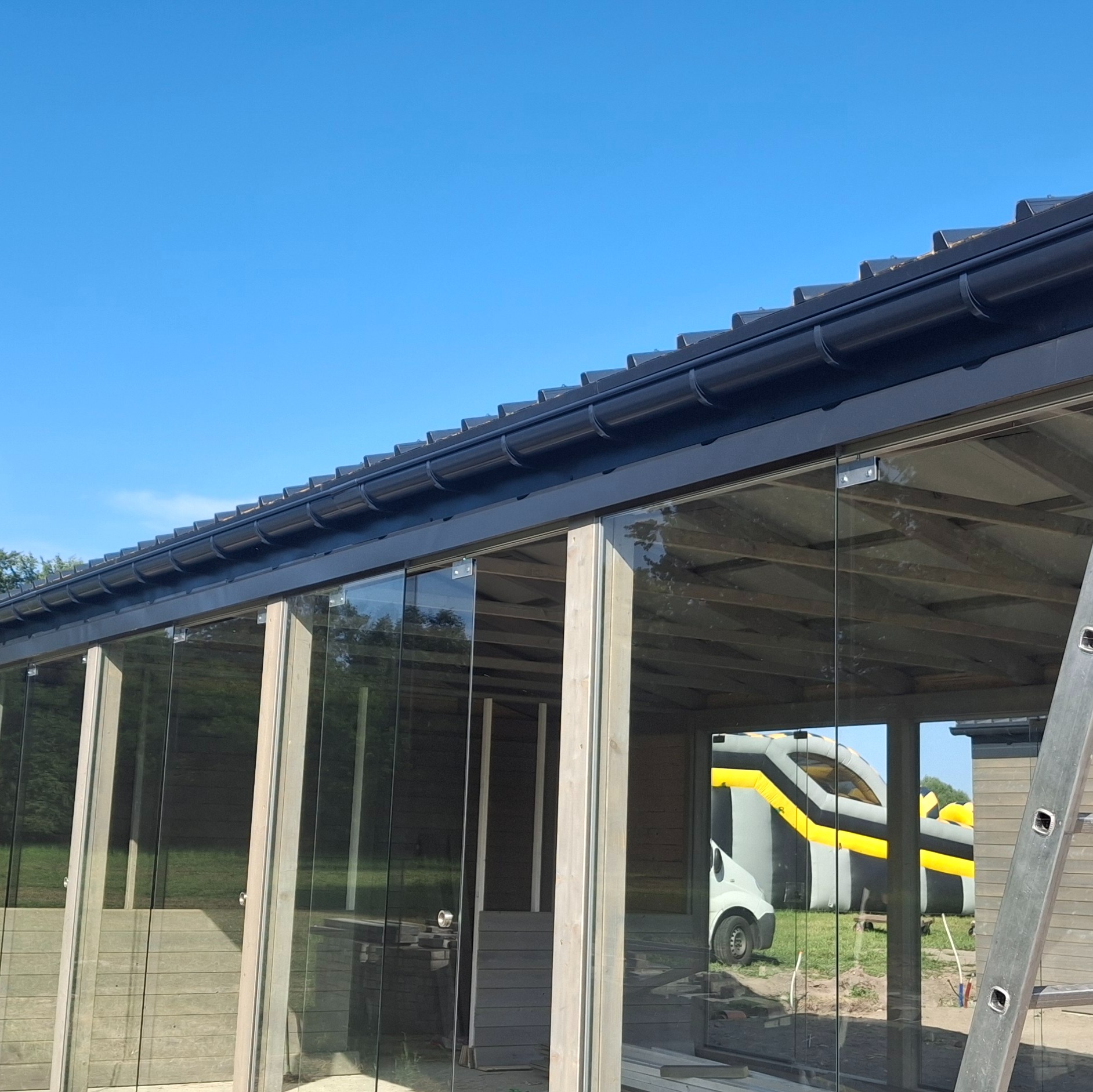 Nowoczesna zabudowa tarasu z przesuwnymi szklanymi panelami i czarnymi rynnami na tle błękitnego nieba. Widoczna drabina i dmuchany zamek w odbiciu szkła.