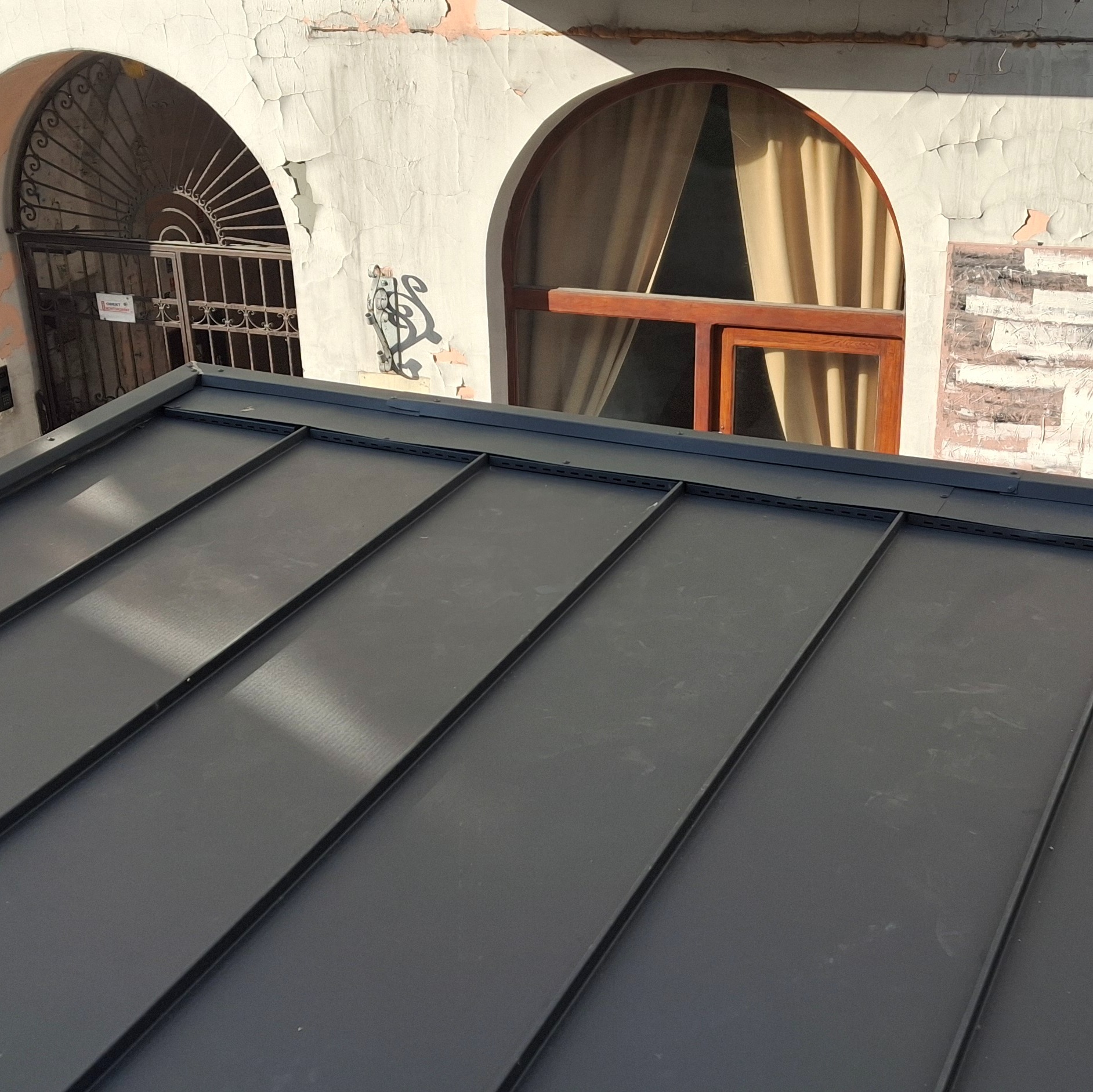 Nowy, grafitowy dach panelowy na tle starej kamienicy z oknami łukowymi i dekoracyjnymi elementami. Widok z góry, podkreślający nowoczesność dachu i kontrast z otoczeniem.