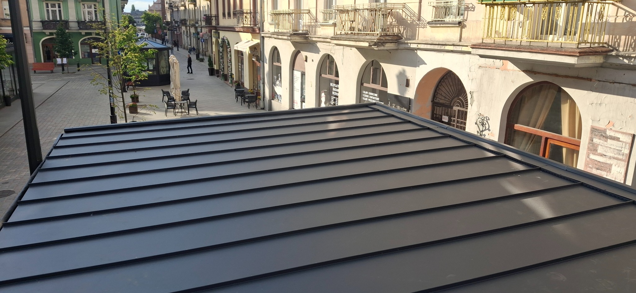 Czarny, metalowy dach na tle miejskiej zabudowy z arkadami i kawiarnianymi stolikami. Widok z góry, podkreślający regularność linii pokrycia dachowego.