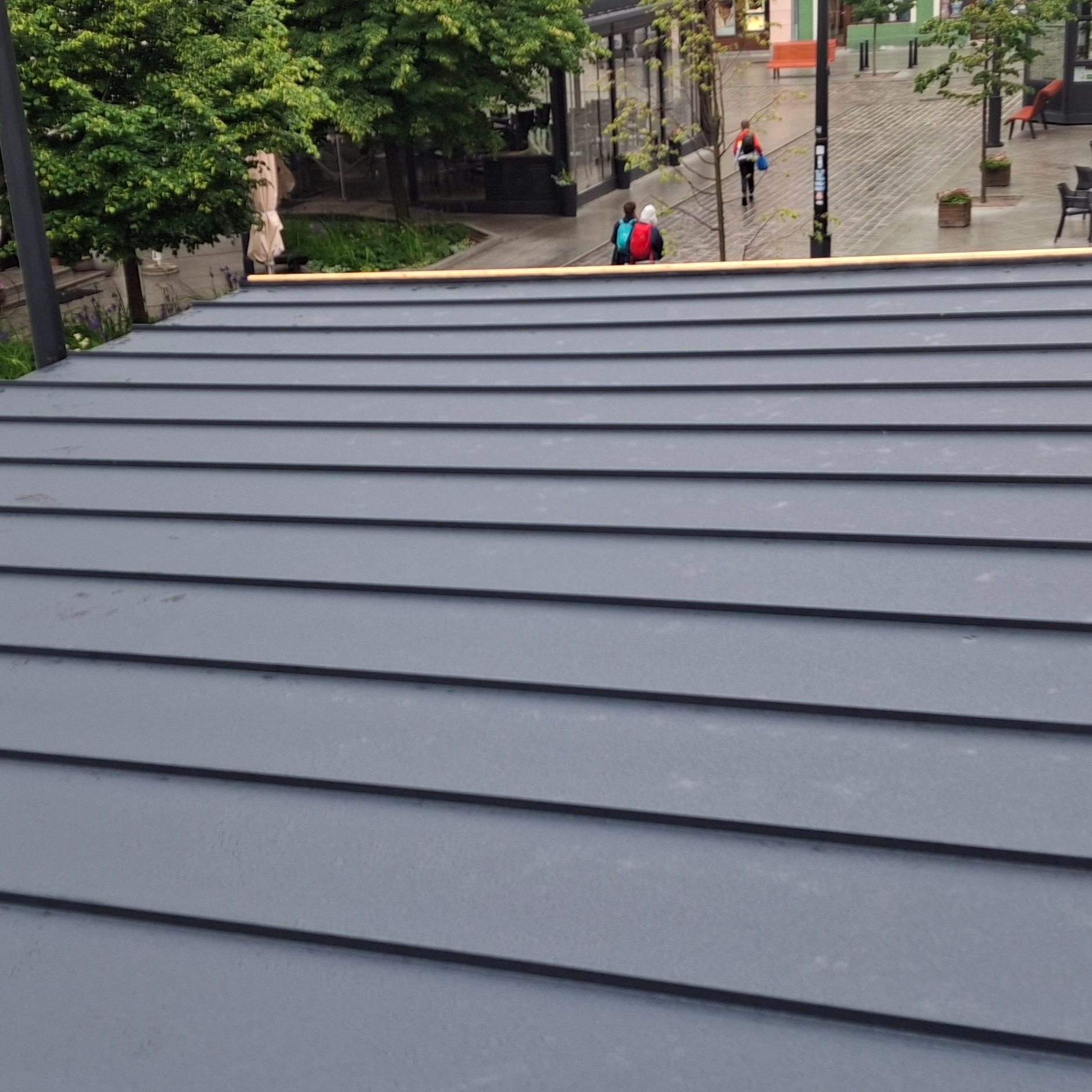 Szary dach z blachy na tle miejskiego placu z zielenią i przechodniami. Widok z góry, prezentujący regularne ułożenie paneli dachowych.