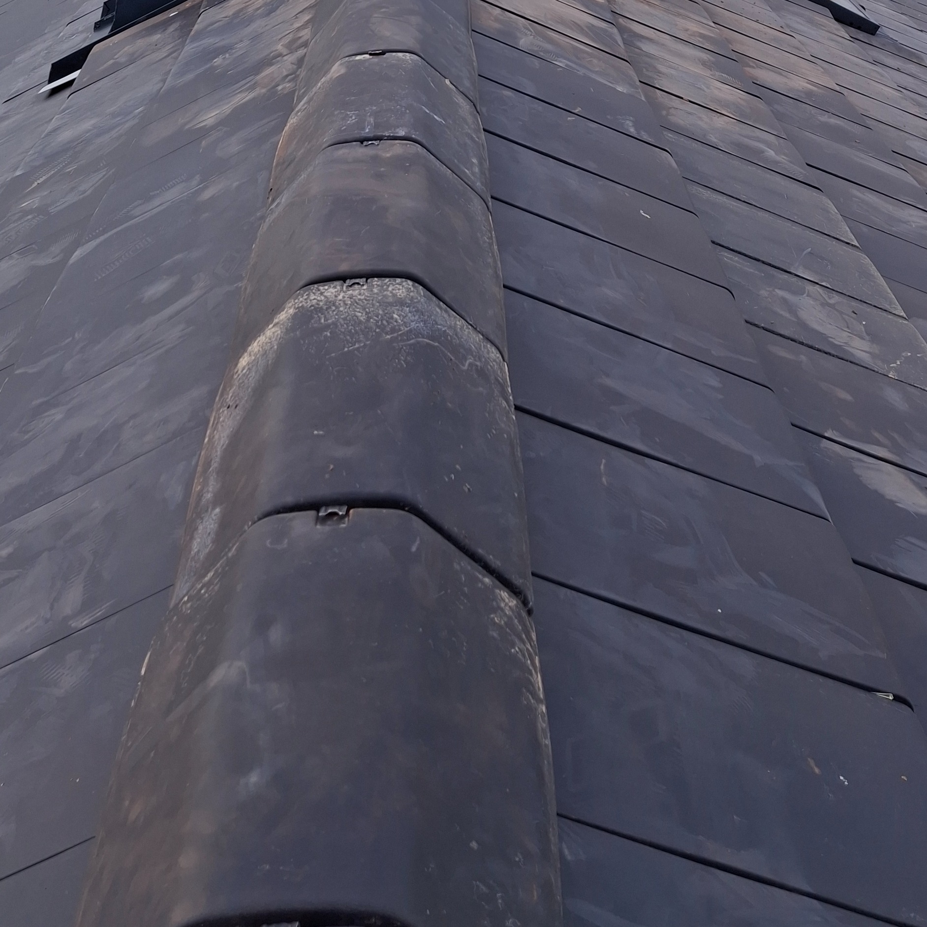 Szczegółowe ujęcie ciemnej, ceramicznej dachówki karpiówki na dachu, z widocznymi elementami mocującymi i drobnymi zabrudzeniami. Układ regularny, perspektywa z dołu.