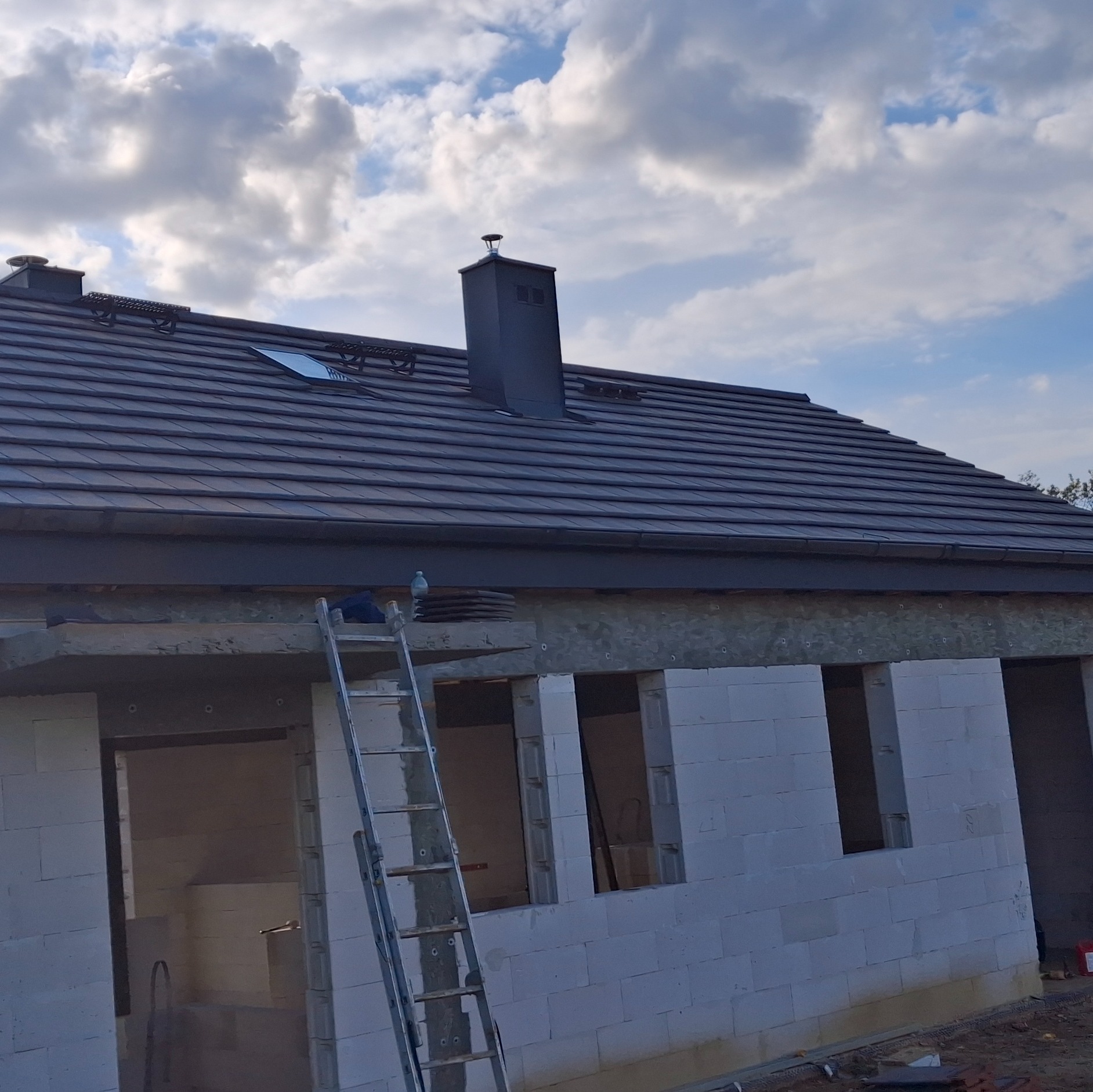 Dachówka ceramiczna na nowym budynku w trakcie budowy, widoczny czarny komin, okno dachowe i drabina oparta o ścianę z betonu komórkowego. Pochmurne niebo w tle.