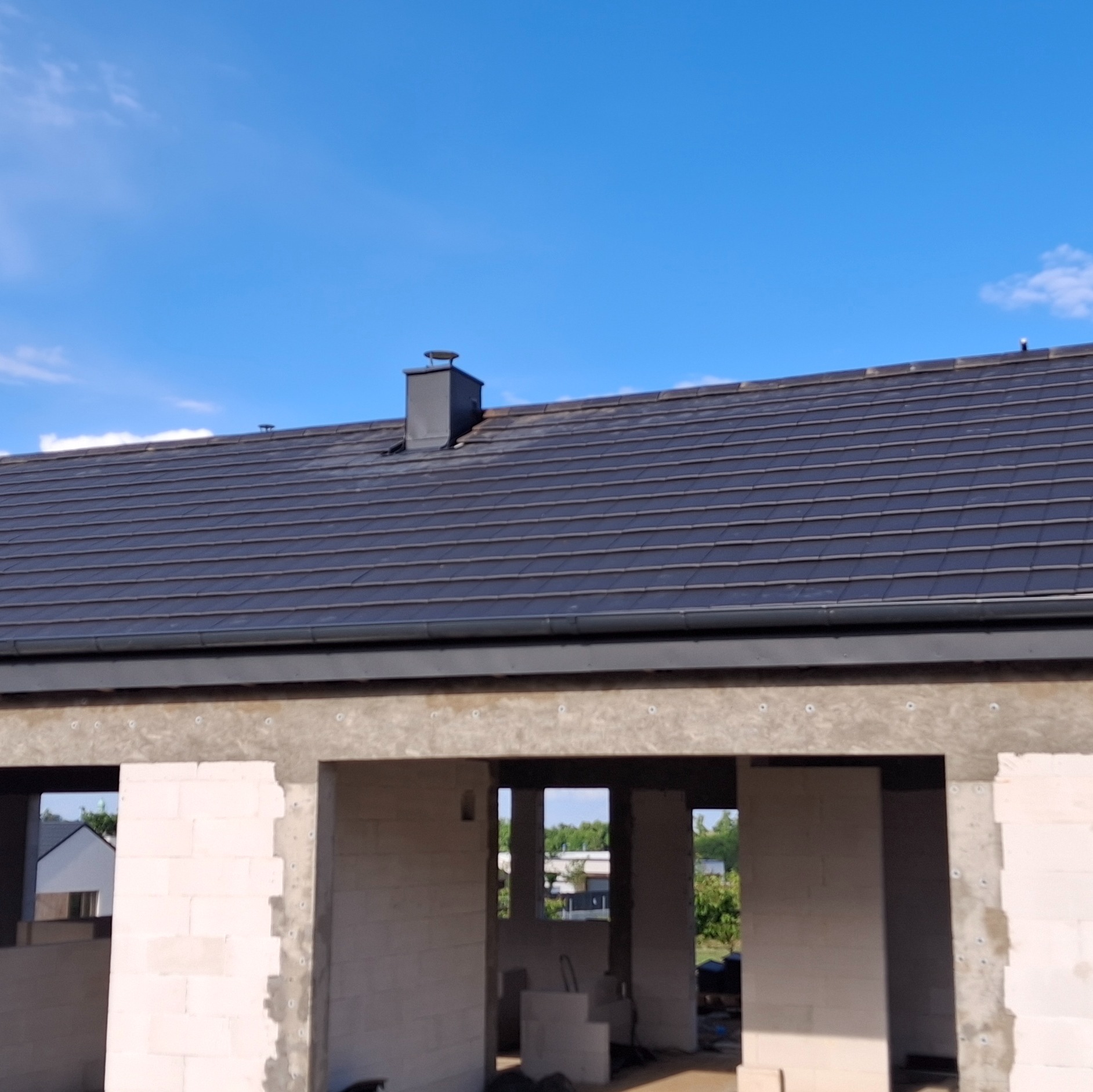 Nowy dach z ciemnej dachówki ceramicznej i obróbki blacharskie na surowym budynku z betonu komórkowego w słoneczny dzień. Widoczne rynny.