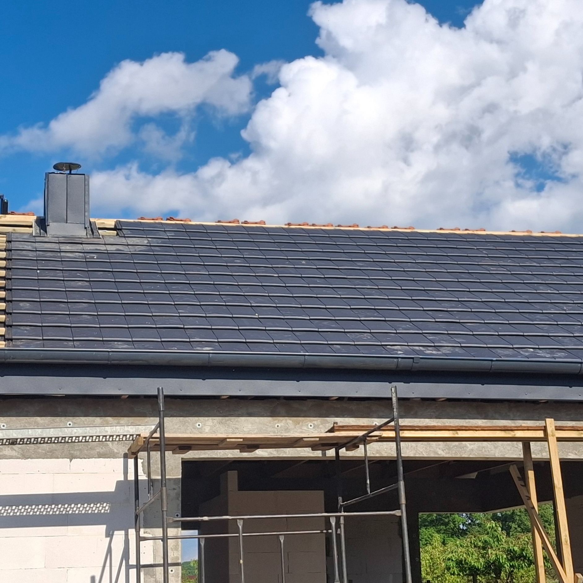 Ciemnoszara dachówka ceramiczna na nowym dachu, z widocznym kominem i fragmentem rusztowania. Błękitne niebo z chmurami w tle.