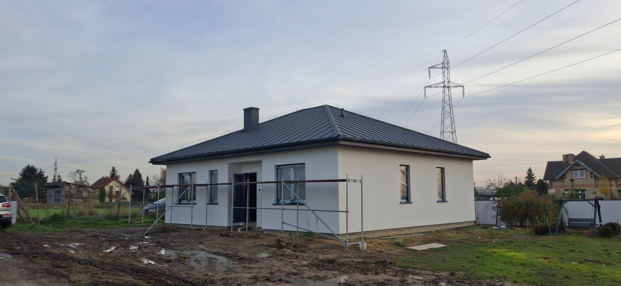 Nowoczesny dom jednorodzinny z ciemnoszarym dachem z blachy na rąbek stojący, białą elewacją i widocznymi rusztowaniami. W tle słup wysokiego napięcia.