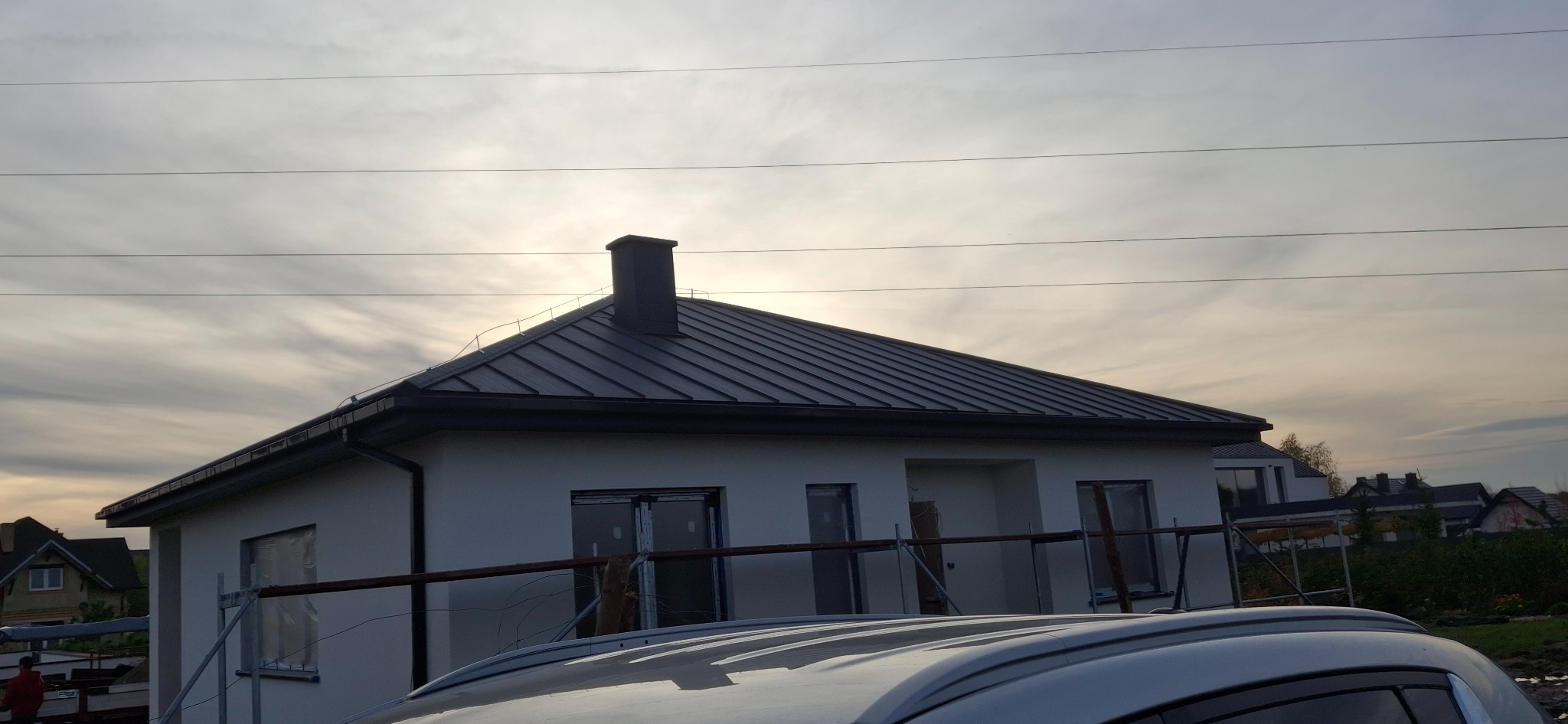 Dom z ciemnoszarym dachem z blachy na tle pochmurnego nieba. Widoczne rusztowania przy ścianie budynku. Ujęcie z dołu, zza samochodu.