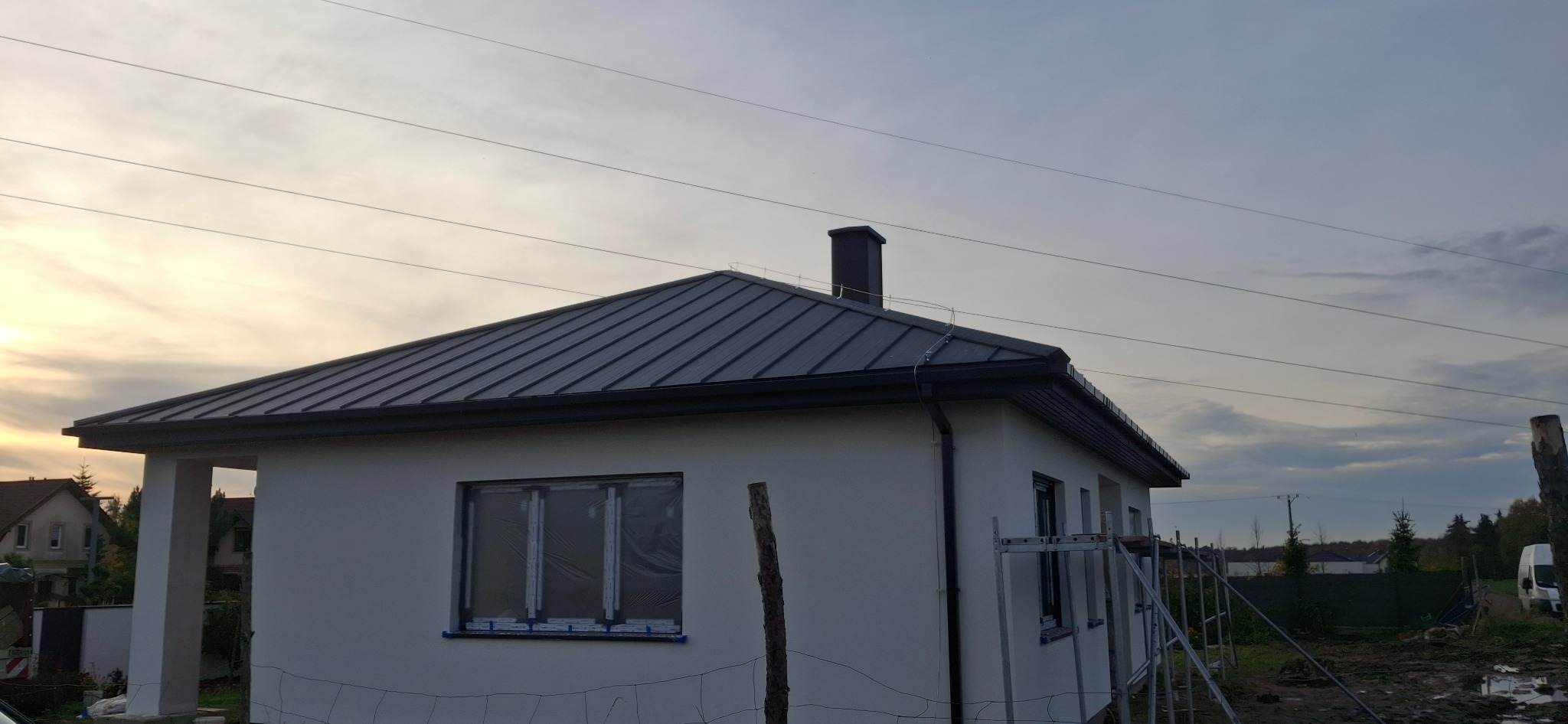 Dom jednorodzinny z ciemnoszarym dachem z blachy na rąbek stojący i orynnowaniem w tym samym kolorze. Okna zabezpieczone folią. Widoczne rusztowanie.