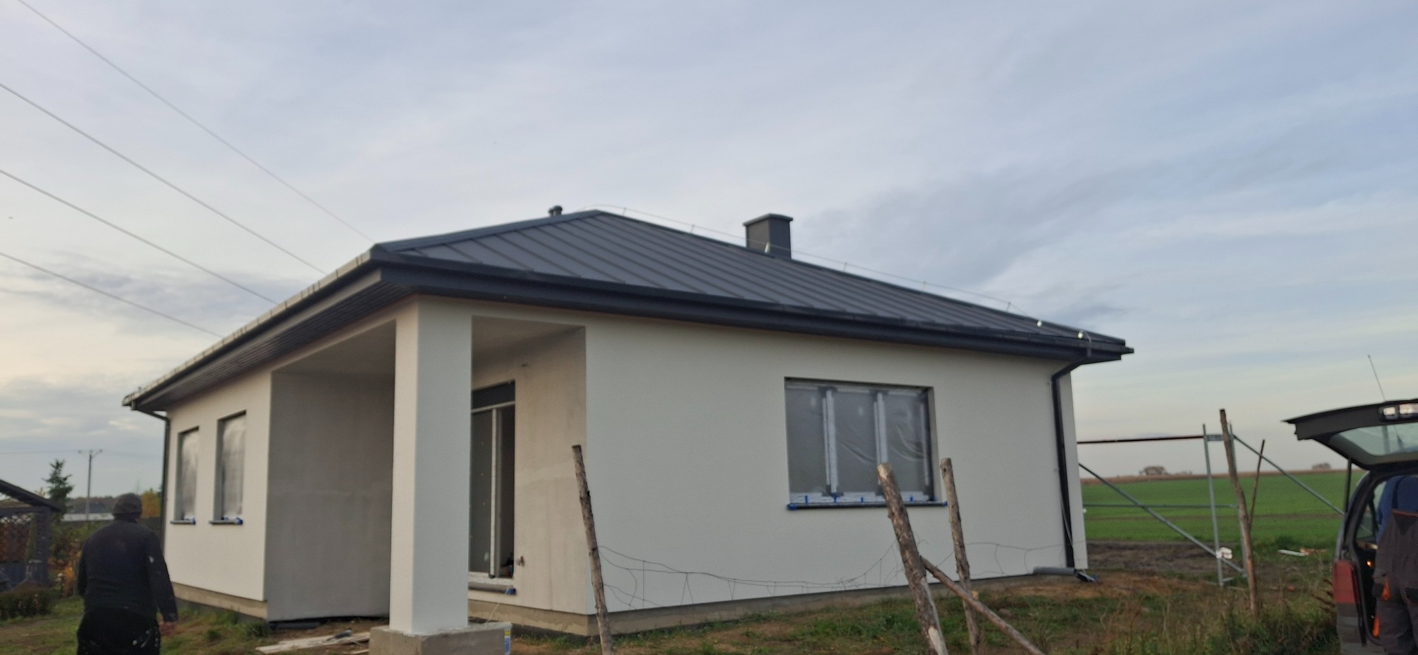 Dom jednorodzinny z ciemnym dachem i jasną elewacją, okna zabezpieczone folią. Widoczne rusztowania i osoby sugerujące prace wykończeniowe.