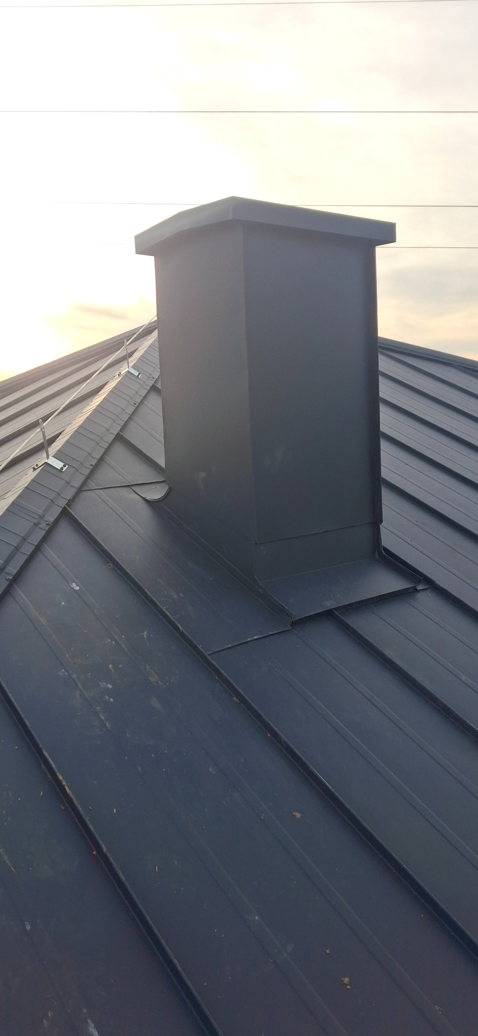 Ciemnoszary, metalowy komin na dachu z blachy na rąbek stojący, zabezpieczony przed zerwaniem. Widoczne mocowania linek. Ujęcie z dołu, w tle niebo.