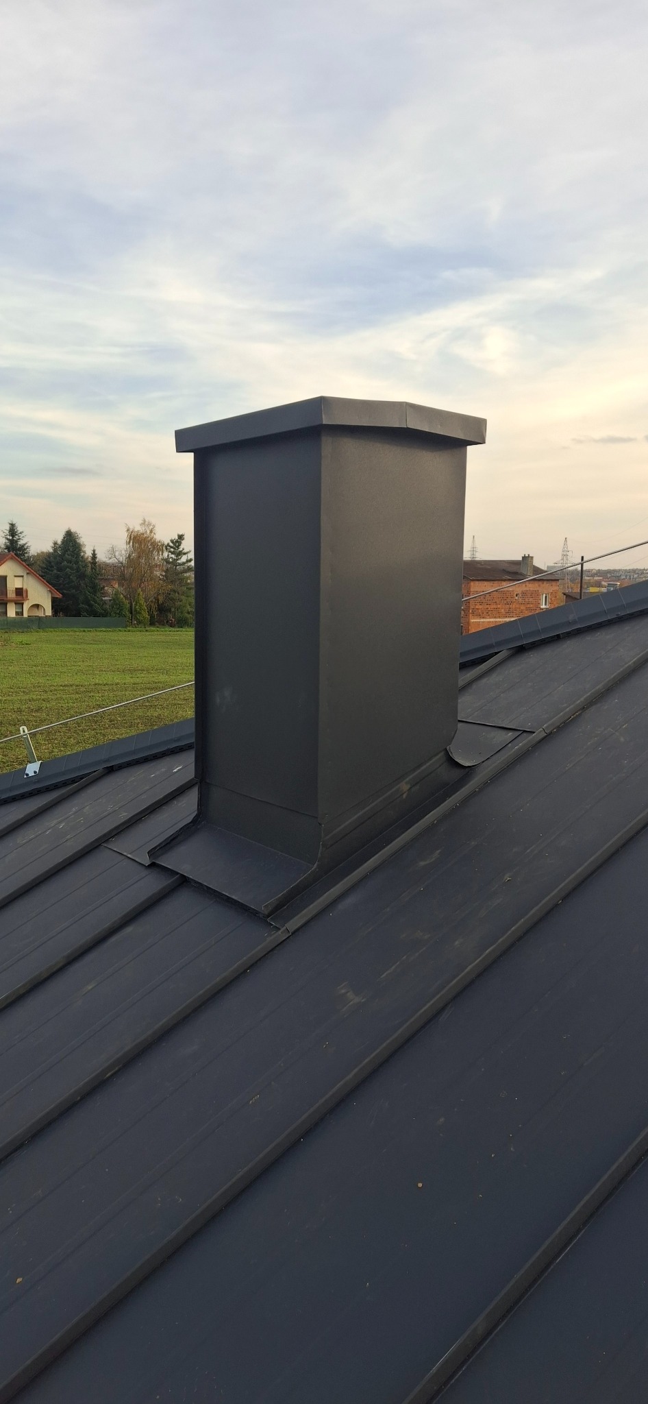 Czarny, metalowy komin na dachu z blachy na tle zieleni i błękitnego nieba. Widoczne obróbki blacharskie.
