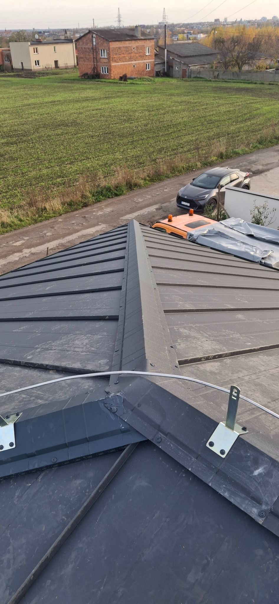 Szary dach z blachy na budynku jednorodzinnym, widok z góry, z elementami montażowymi i zabezpieczeniami. W tle pole i zabudowania.