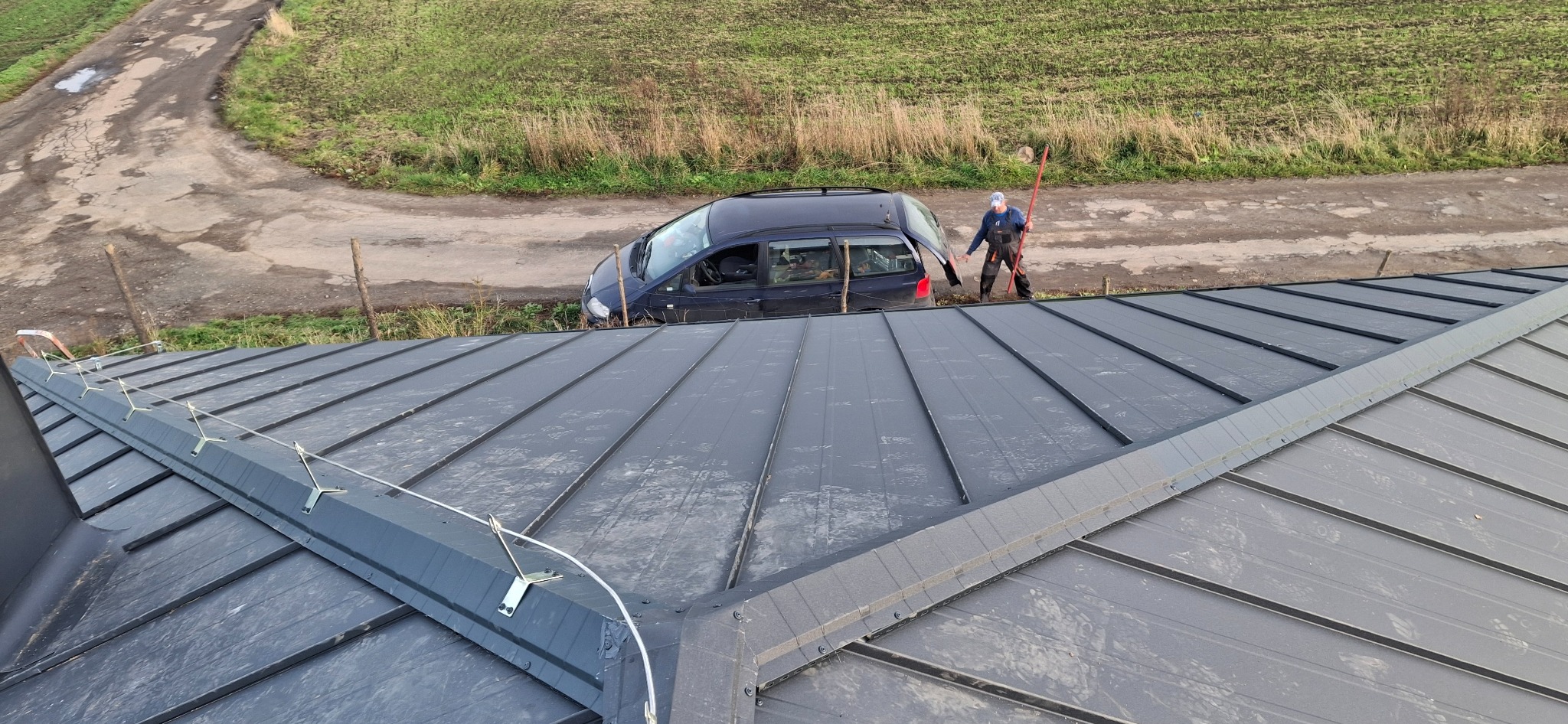 Szary dach z blachy na rąbek stojący, zabezpieczony systemem odśnieżania. W tle auto i dekarz z narzędziem na tle pola i drogi.
