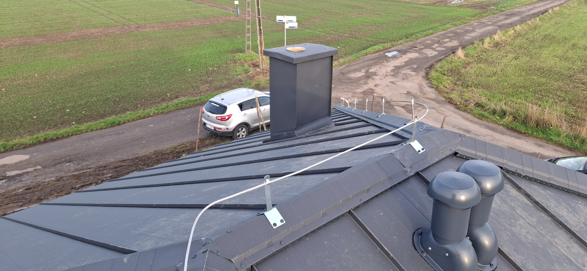 Grafitowy dach z blachy na rąbek stojący, z kominem i odpowietrznikami. Widoczne zabezpieczenia przeciwśniegowe i auto na drodze gruntowej w tle.