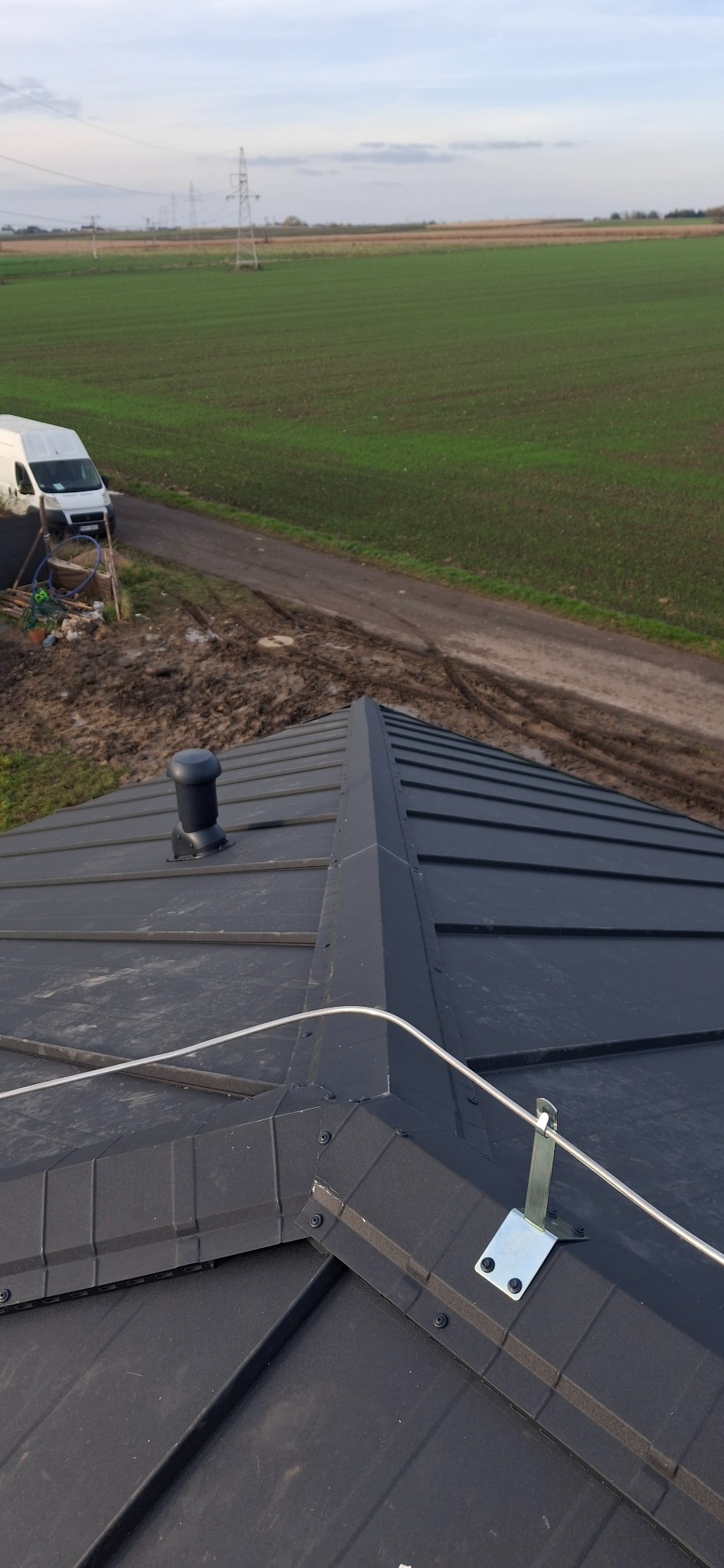 Czarny dach z blachy na domu jednorodzinnym, widok z góry, z zamontowanym odpowietrznikiem i zabezpieczeniem przeciwśniegowym. W tle pole i zaparkowany bus.