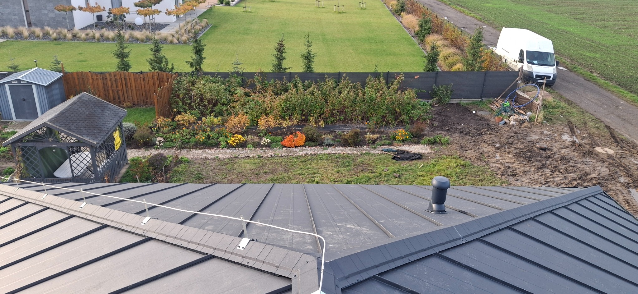 Grafitowy dach z blachy na tle zadbanego ogrodu i białego busa. Widoczny system odśnieżania. Ujęcie z góry podkreśla geometrię dachu i estetykę otoczenia.