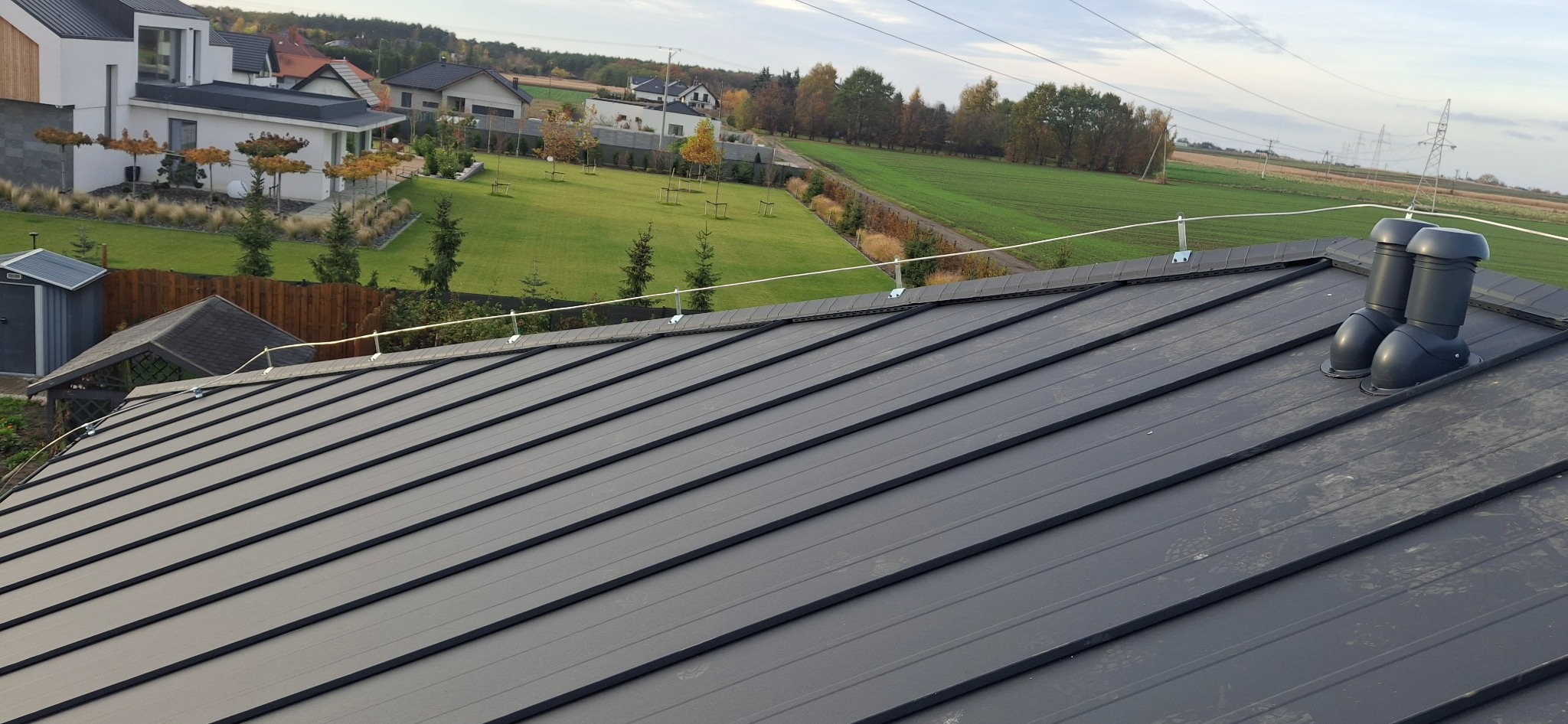 Grafitowy dach z blachy na tle zielonego krajobrazu. Widoczne kominki wentylacyjne i system odgromowy. Ujęcie z góry, podkreślające regularność wzoru blachy.