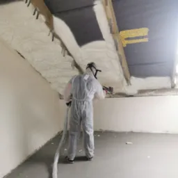 Pracownik w kombinezonie ochronnym aplikuje piankę izolacyjną na strop poddasza, widoczne elementy konstrukcyjne dachu i mocowania, szara podłoga w surowym stanie.