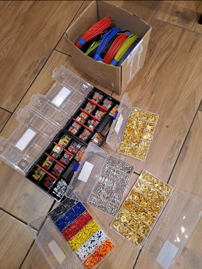 Zestaw różnokolorowych przewodów w kartonie oraz plastikowe pojemniki z konektorami i złączkami elektrycznymi na drewnianej podłodze.